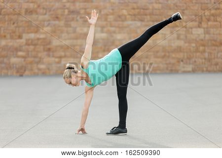 Sporty beautiful young woman practicing yoga, standing in Half Moon exercise, Ardha Chandrasana pose, working out, wearing sportswear, outdoor full length, brick wall background