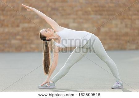 Sporty attractive young woman practicing yoga, standing in Extended triangle exercise, Utthita Trikonasana pose, working out, wearing sportswear, outdoor full length, brick wall background