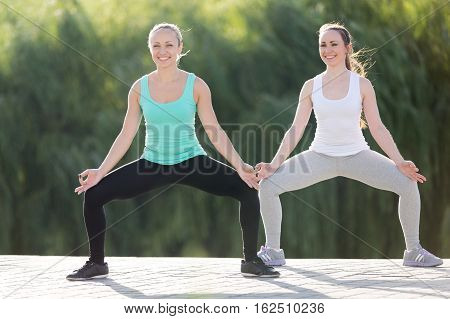 Group of two cheerful sporty attractive young women practicing yoga, standing in Sumo Squat exercise, Goddess pose, working out, wearing sportswear, outdoor full length, street background