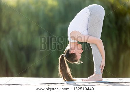 Sporty smiling attractive young woman practicing yoga, doing standing forward bend, head to knees exercise, uttanasana pose, working out, wearing sportswear, outdoor full length, street background