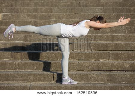 Sporty attractive young woman practicing yoga, standing in Warrior Three exercise, Virabhadrasana 3 pose, working out, wearing sportswear, outdoor, stone stair background with a shadow