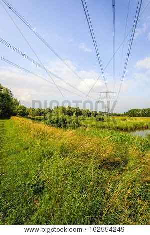 High voltage lines and pylons in a large nature reserve with small creeks in the Netherlands. It's a sunny day in the summer season.