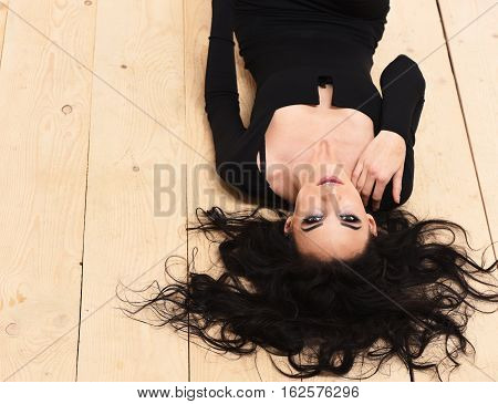 pretty cute sexy girl or beautiful young woman with fashion makeup on serious face and curly long brunette hair in black dress with deep neckline lying upside down on wooden floor background