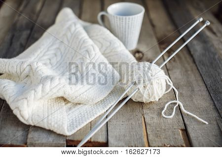 Beautiful white knitted scarf and ball of wool with needles and cup of tea on old wooden table. Cozy winter. Hobbies. Selective focus.
