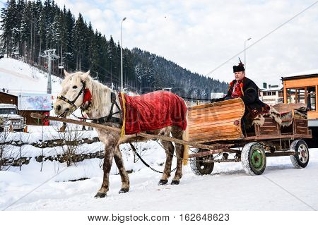 BUKOVEL, UKRAINE - December 17, 2016: Wooden cart with white horse and man in ukrainian national suite of ski resort Bukovel, Ukraine. Bukovel is the most popular ski resort in Ukraine.Carpathian.