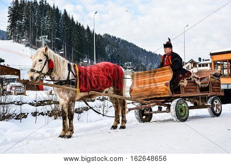 BUKOVEL, UKRAINE - December 17, 2016: Wooden cart with white horse and man in ukrainian national suite of ski resort Bukovel, Ukraine. Bukovel is the most popular ski resort in Ukraine.Carpathian.