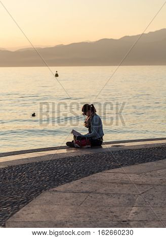 LAZISE ITALY - MAY 5 2016: Young woman reading a book with a sunset view on the promenade of Lake Garda in Lazise Italy