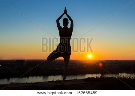 Silhouette of sporty woman practicing yoga in the park at sunset - tree pose, vrikshasana. Sunset light, golden hour. Freedom, health and yoga concept