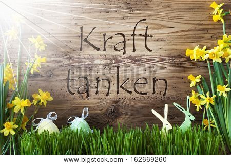 Wooden Background With German Text Kraft Tanken Means Relax. Easter Decoration Like Easter Eggs And Easter Bunny. Sunny Yellow Spring Flower Narcisssus With Gras. Card For Seasons Greetings