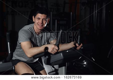 Athletic Man Smiling On Excersice Bike At The Gym And Looking At Camera.