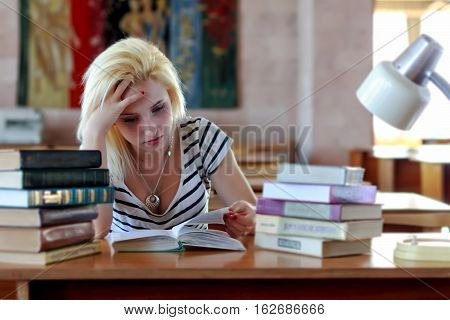 Tired blond, student or schoolgirl, looking thoughtfully into the book while sitting at a desk