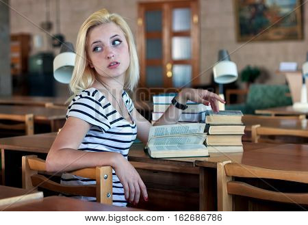 Tired girl coquettishly turns around to look back. She sits at a table on which there are a pile of books and reading lamps