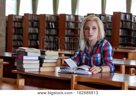 Young blond girl in a plaid shirt sitting at a table in the old library with the book in his hands. On the table are piles of books, visible in the background of bookshelves