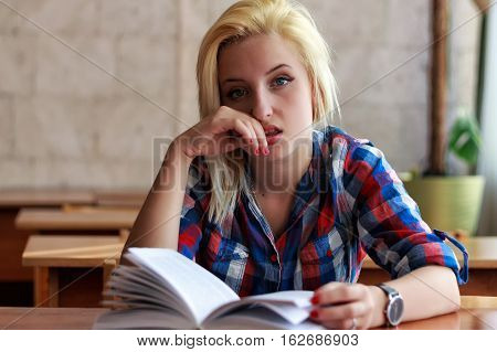 Dissatisfied blond girl in plaid shirt with an open book sitting at a desk in the classroom