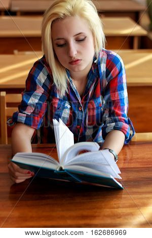 Young blonde in a plaid shirt sitting at the Desk in the classroom, holding an open book