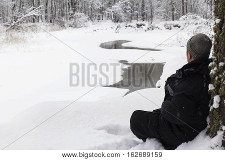 a young man sitting in the snow snowdrift in the forest and looking at the winter river