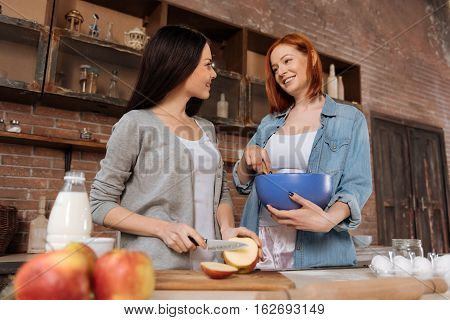 Happy time. Same sex couple wearing casual clothes standing in semi position near the table mixing and cutting apple