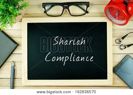 Top view of earphone, calculator, alarm clock, spectacle, notebook, pen, smartphone and chalkboard written with SHARIAH COMPLIANCE.