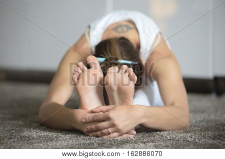 Sporty young woman practicing yoga, seated forward bend, exercise, paschimottanasana pose, working out, wearing white sportswear, indoor, home interior background, closeup. Focus on feet