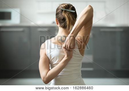 Close up of female hands behind the back, woman practicing yoga, sitting in Cow Face exercise, Gomukasana pose, working out, wearing white sportswear, indoor, home interior background