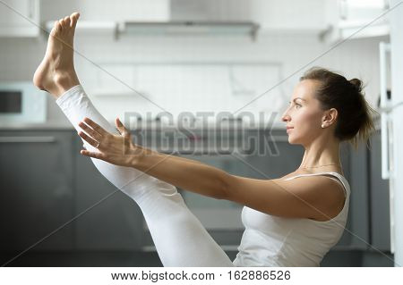 Side view portrait of sporty attractive woman practicing yoga, sitting in balance exercise, Paripurna Navasana pose, working out, wearing white sportswear, indoor, home interior background