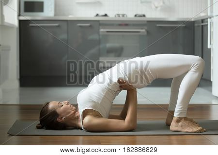 Sporty attractive woman practicing yoga, stretching in Bridge exercise, dvi pada pithasana pose, working out, wearing white sportswear, indoor full length, home interior background