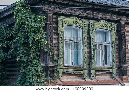 Old carved windows with frames, the wall of a wooden house covered with vines and ivy