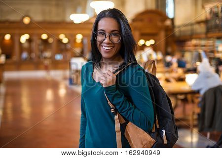 Happy college student girl with backpack in the library smiling. Beautiful young student woman in reading room. Education smile study learning