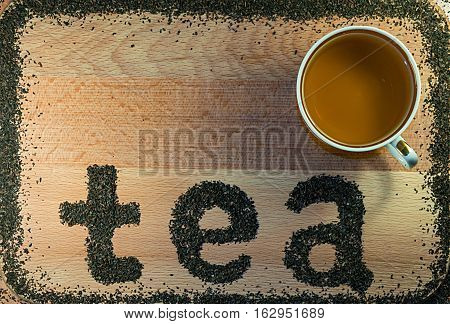 The word tea is laid out on a light-brown wooden Board small leaf black tea. Board is the background. The edges lined with a frame of tea. On Board is a mug.