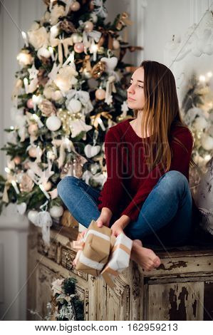 Woman with a Christmas Gift sitting on acncient commode decorated for xmas. Fir tree at background.