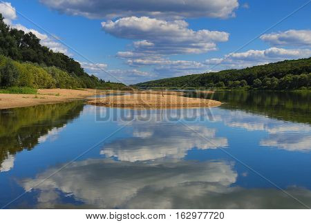 view of the water in which is reflected the blue sky and clouds and a sandy island