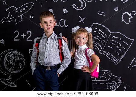 School Mates, Boy And Girl, Standing Before The Chalkboard As A Background With A Backpack On Their 