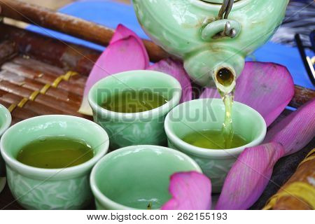 Pouring Lotus Tea From A Porcelain Teapot Into Celadon Glazed Cups On A Bamboo Tray With Pink Lotus 
