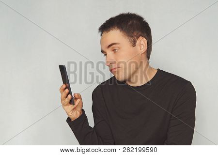 A Young Guy Looks At The Screen Of A Black Smartphone With A Dismissive Emotion On His Face. Uninter