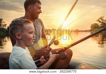 Happy Father and Son together fishing from a boat at sunset time in summer day under beautiful sky on the lake.