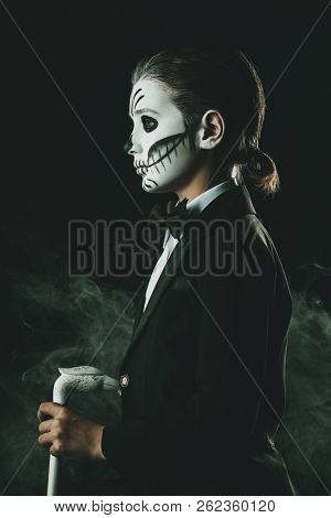 Portrait of a child boy with carnival make-up of a skeleton wearing elegant tail-coat. Studio shot over black background. Halloween. Dia de los muertos. The Day of the Dead.