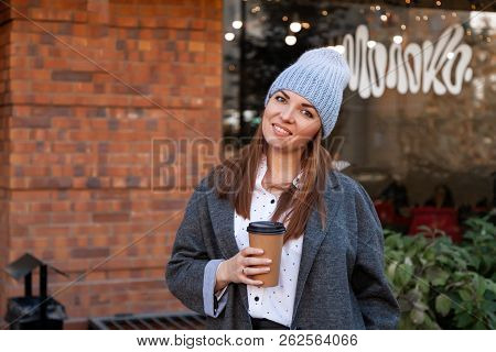 Young Attractive Lady In Gray Knitting Hat And Gray Coat Drink Coffee To Go . Smiling Beautiful Hips