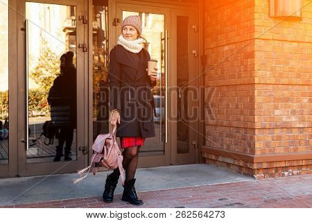 Young Attractive Lady In Brown Knitting Hat And Black Coat Drink Coffee To Go . Smiling Beautiful Hi