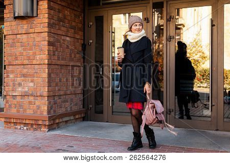 Young Attractive Lady In Brown Knitting Hat And Black Coat Drink Coffee To Go . Smiling Beautiful Hi