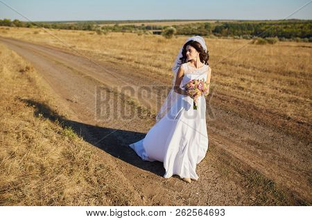 Beautiful Smiling Bride Wearing A Natural Flower Wreath And Standing In The Golden Summer Fields Wit