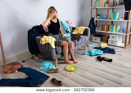 Upset Woman Sitting On Sofa Surrounded By Scatted Clothes