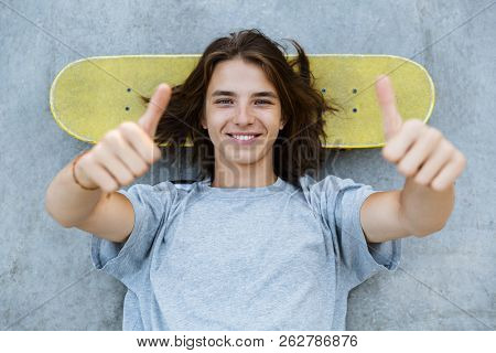 Top view of a cute young teenge boy spending time at the skate park, laying on a skateboard at a ramp, showing thumbs up