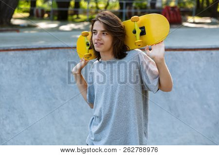 Smiling young teenge boy spending time at the skate park, holding a skateboard, looking away