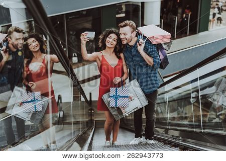 Couple With Packages Taking Selfie On Escalator. Using Smartphone. Digital Device In Hand. Shopping 