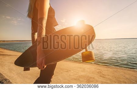 A Beautiful, Sexy Hipster Girl In Tattoo Stands With A Longboard Against The Sunset At The Sea. Long