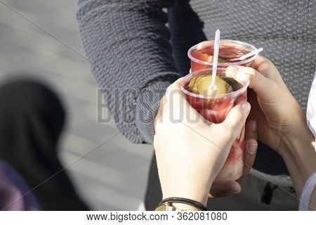Gravity Of Female And Male Hands. The Cocktail Is Pickled, They Hold Plastic Cups And There Is A Pla