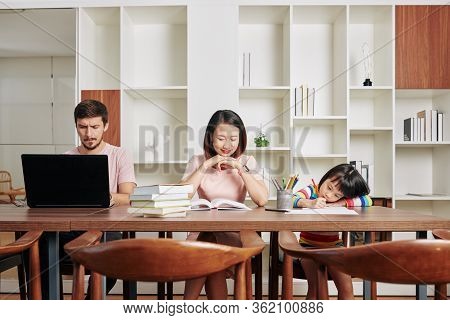 Little Asian Girl Drawing Picture, Mother Reading A Book And Father Working On Laptop At Home