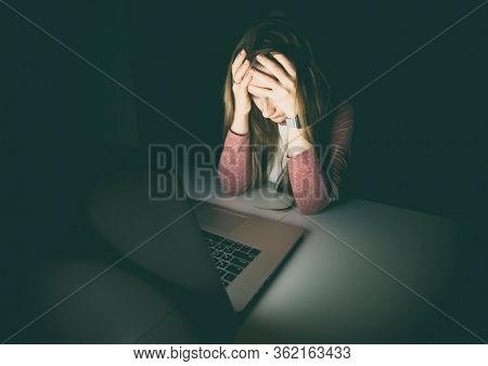 Very tired and exhausted woman, holds her head with hands sitting at the laptop  late in the night. Concept of the workaholic, overworking and anxiety