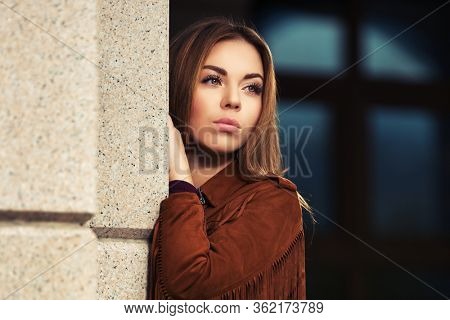 Sad young fashion woman leaning on the wall Stylish female model in brown leather fringe suede jacket