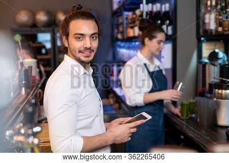 Young friendly waiter in white shirt scrolling through online menu while checking prices for drinks and food on background of colleague